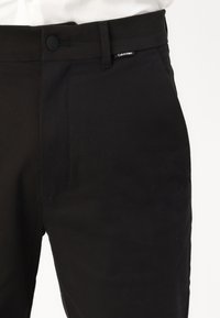 Pantaloni negri dintr-un material cu textură netedă. Au închidere cu buton, un buzunar lateral și un mic etichetă Calvin Klein pe talie.