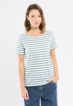Femme aux cheveux bruns mi-longs portant un t-shirt à manches courtes blanc à rayures vertes et un jean bleu, debout avec une main dans la poche.