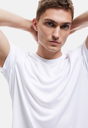 Ung mand med kort hår iført en hvid t-shirt, med løftede arme og hænder hvilende bag hovedet, kigger fremad.