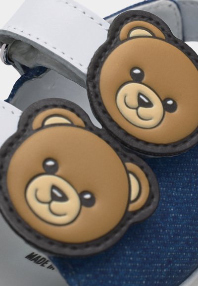 Παιδικό παπούτσι με μπλε ύφασμα, λευκό λουράκι και δύο διακοσμήσεις με πρόσωπα καφέ αρκούδας στο λουράκι.