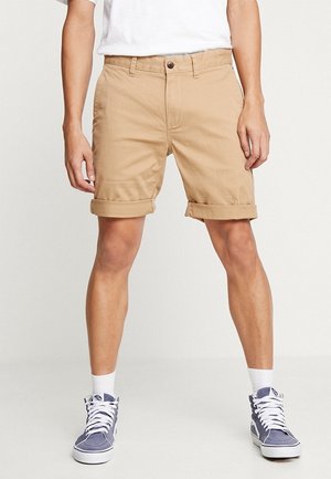 Khaki shorts met opgerolde zoom, gemaakt van katoen, met een voorste knoopsluiting en zijzakken. Draagt bij blauwe hoge sneakers.