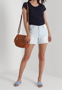Kobieta w granatowej koszulce, jasnych, podwiniętych krótkich spodenkach dżinsowych, niebieskich klapkach oraz z brązową torebką na ramię z kieszenią na zamek.