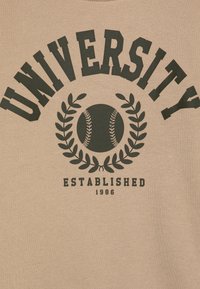 Beige pulóver, amelyen vastag fekete betűkkel áll a "UNIVERSITY" felirat egy baseball és babérkoszorú grafika felett, és az "ESTABLISHED 1986" felirat alatta.