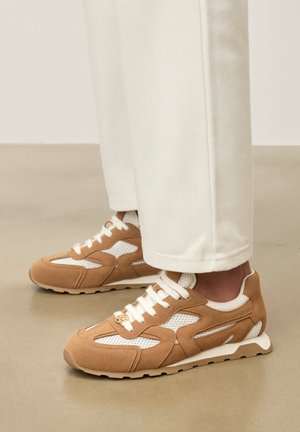 Pruunid ja valged võrgust tossud koos heledate pükstega, seisavad siledal beežil põrandal.