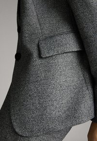 Blazer gris texturé avec un revers cranté, une fermeture à un bouton et une poche poitrine. Tissu lisse avec un motif subtil.