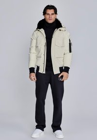 Giubbotto imbottito di colore crema chiaro con dettagli neri, dotato di molteplici tasche frontali e un cappuccio. Abbinato a pantaloni tuta neri e sneakers bianche.