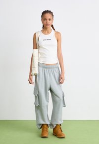 Hvid ribbet tanktop med "Dickies" logo, grå cargo sweatpants med sidelommer og brune ankelstøvler. Simpel tekstildesign.