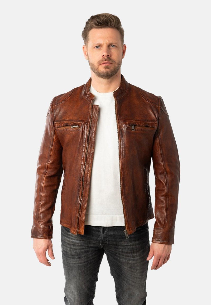 Chaqueta de cuero marrón con cierre frontal de cremallera, cuello redondo y dos bolsillos en el pecho con cremallera. Lleva puesta sobre una camisa blanca y combinada con vaqueros grises.