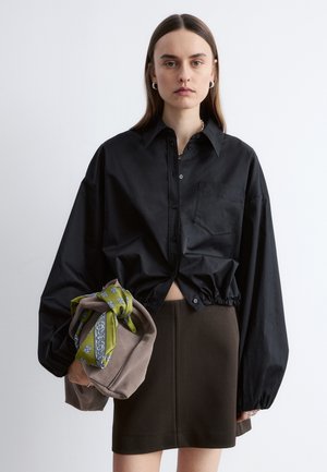 Vrouw met lang bruin haar draagt een oversized zwart overhemd en een donkere rok, en houdt een taupekleurige handtas vast waaraan een groen sjaaltje met patroon is geknoopt.