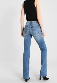 Vaqueros de mezclilla azul claro de corte acampanado con un acabado desvaído, bolsillos traseros y una etiqueta de cuero marrón en la cintura. La modelo lleva una camiseta negra sin mangas.
