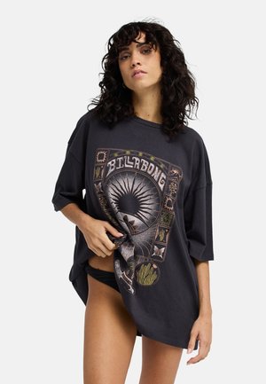 Femme aux cheveux bouclés portant un t-shirt Billabong noir oversize avec des motifs soleil et nature, tenant l'ourlet pour révéler un bas de bikini noir.