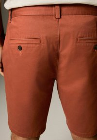 Shorts de culoare ruginie, din bumbac, cu o textură netedă, având buzunare la spate cu butoane negre și un croi drept.