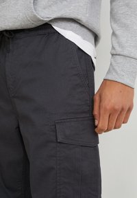 Mörkgrå cargo byxor med en smal passform, med sidoficka och elastisk midja med dragsko. Tyget har en slät yta.