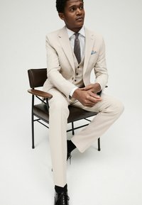 Costume beige en trois pièces avec une coupe ajustée, un revers cranté et trois boutons. Porté avec une chemise blanche, une cravate à pois et des chaussures noires.