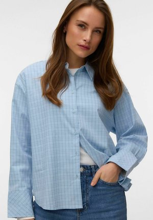 Femeie cu păr lung și castaniu, purtând o cămașă în carouri albastru deschis peste un tricou alb și blugi albaștri, cu mâna în buzunar.