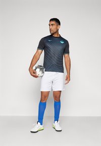 Camiseta deportiva negra con acentos azules y patrón de ondas, pantalones cortos blancos, calcetines azules y zapatos blancos, sosteniendo un balón de fútbol en blanco y negro.