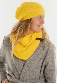 Geel gebreide beanie en bijpassende infinity-sjaal, beide met een gestructureerd patroon, gelaagd over een lichtgrijs gestructureerd jasje. Accentueert met bruin leer.