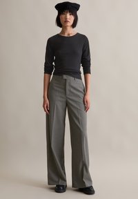 Schwarzes Langarmshirt kombiniert mit grauen, gestreiften, weiten Hosen. Das Outfit zeichnet sich durch eine hohe Taille, Vorderfalten und klobige schwarze Schuhe aus.