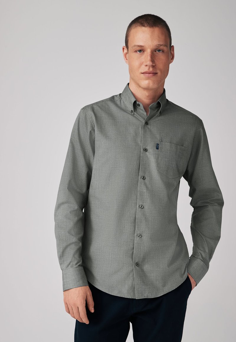 Man wearing a long-sleeve gray button-up shirt with a chest pocket, standing against a plain light gray background.