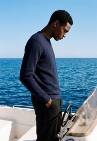 Homme en pull bleu marine et pantalon noir debout sur un bateau, regardant le volant, avec l’océan et un ciel dégagé en arrière-plan.