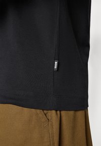 Zwart katoenen T-shirt met een zijlabel met de merknaam "BOSS", gecombineerd met bruine stoffen broek, met een casual outfitstijl.