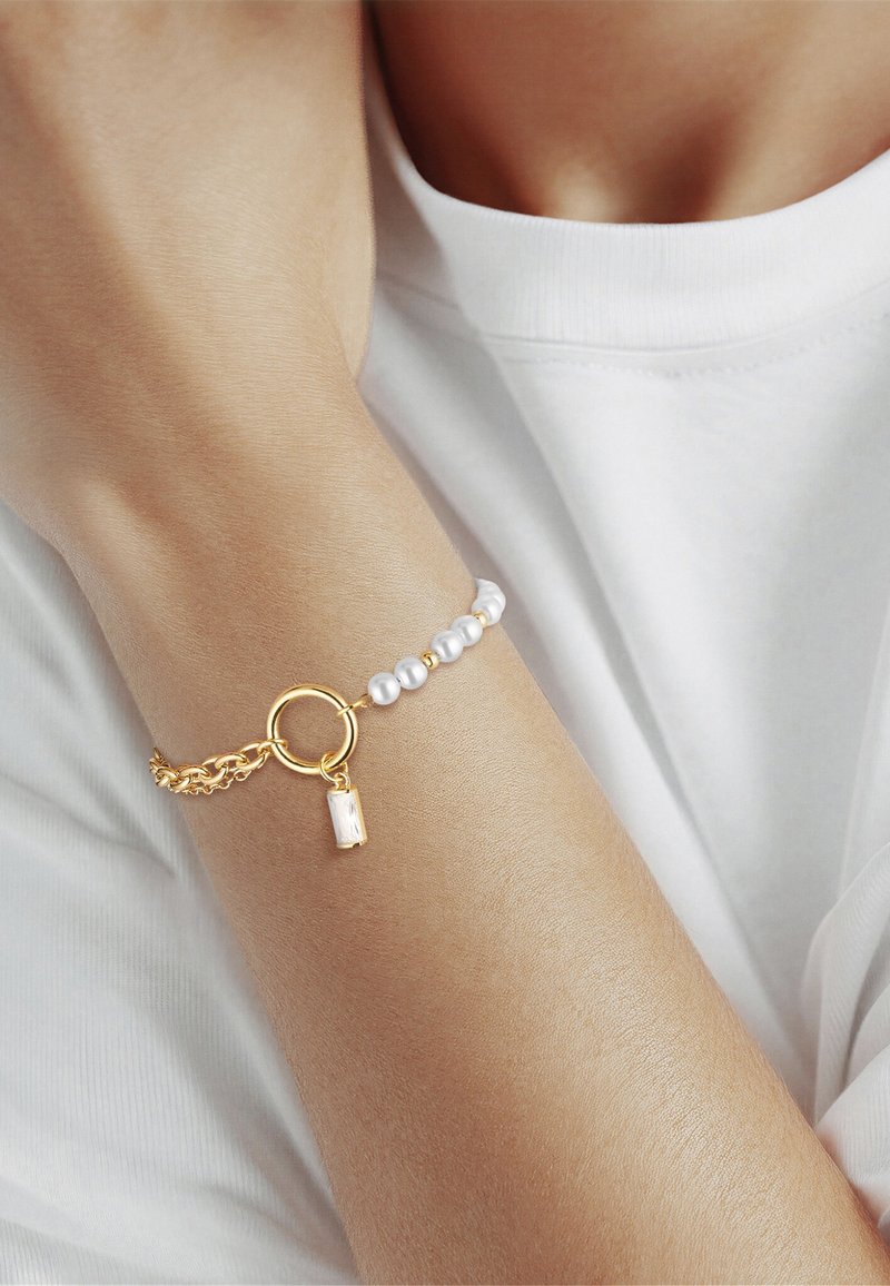 Handgelenk mit einem Armband mit weißen Perlen und goldenen Perlen, einem goldenen runden Verschluss und einem kleinen rechteckigen Anhänger, getragen über einem weißen Hemd.