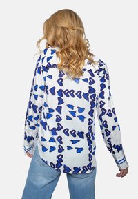 Biała koszula z długimi rękawami w niebieskie serduszka, cechująca się luźnym krojem i mankietami z guzikami, zestawiona z jasnoniebieskimi dżinsami.