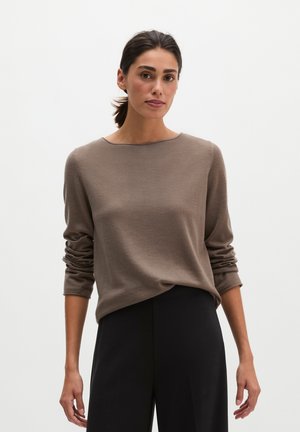Kobieta o ciemnych włosach związanych w kucyk, ubrana w taupe'owy sweter z długim rękawem i czarne spodnie na jednolitym jasnym tle.
