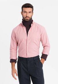 Lichtroze overhemd met lange mouwen, gecombineerd met een donker turtleneck en donkergrijze broek. Heeft een borstzak en opgerolde mouwen.