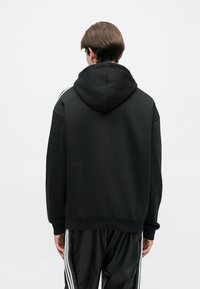 Čierna mikina s kapucňou a voľným strihom, so strapcami v bielej farbe na pleciach a rebrovanými manžetami. Hladká textúra látky.
