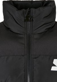 Musta puffer-takki, jossa on korkea kaulus, koko pituinen vetoketju, teksturoitu kangas ja valkoinen logo-aksentti etupuolella.