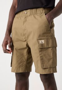 Pantaloni cargo beige con cintura elasticizzata, due tasche laterali e una tasca applicata sulla coscia con dettaglio del logo. Tessuto texture.
