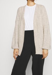 Light beige knitted cardigan with open front, long sleeves, and ribbed texture, paired with a white t-shirt and black high-waisted trousers.