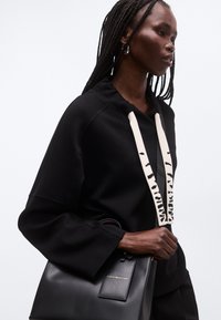 Kvinde med flettet hår iført en sort hættetrøje med hvide snore og holder en sort lædertaske med en guldtags.