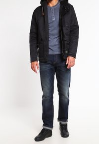 Sort hættetrøje over en grå henley skjorte, parret med mørke denim jeans og sorte sneakers. Har lommer og en afslappet pasform.