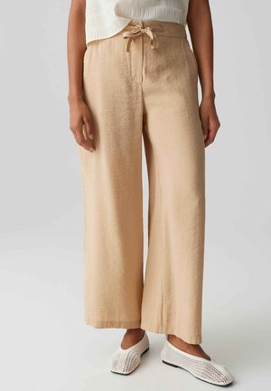 Femme portant un pantalon beige à jambes larges avec une taille nouée et des chaussures blanches en maille ouverte à enfiler, debout sur un fond uni.