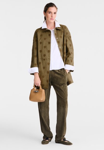 Woman wearing olive green patterned coat, white collared shirt, olive wide pants, holding small tan handbag, and wearing dark flats on white background.