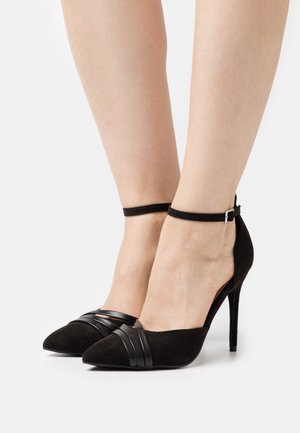 Chaussures à talons hauts en daim noir avec bouts pointus, ornées de détails à brides noires sur le devant et de sangles de cheville avec ferrures argentées.