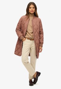 Femme portant un long manteau matelassé mauve par-dessus un col roulé beige, un pantalon crème avec une ceinture tressée et des mocassins marron foncé, se tenant avec une main dans la poche.