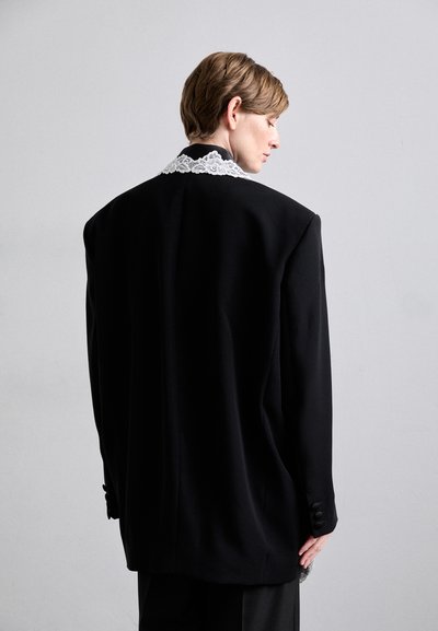 Zwarte oversized blazer met gestructureerde schouders, versierd met een witte kanten kraag en zwarte knoopaccenten op de mouwen.