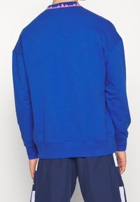 Personne portant un sweat-shirt bleu vif avec un col à motif rose, associé à un short bleu marine à rayures blanches.