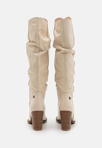 Botas de cuero color crema hasta la rodilla, con una textura ligeramente arrugada, punta redonda y un tacón de madera apilado de 7 cm; cuentan con una cremallera lateral.