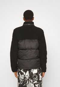 Schwarze Daunenjacke mit Fleece-Oberteil, ausgestattet mit einem hohen Kragen und Knopfverschluss. Strukturierter Stoff kontrastiert mit glattem Nylon.