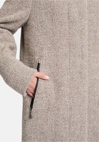 Beige textuur jas met een close-up van een zijzak met een zwarte rits. De stof is zacht en fuzzy, met zichtbare stiksels.