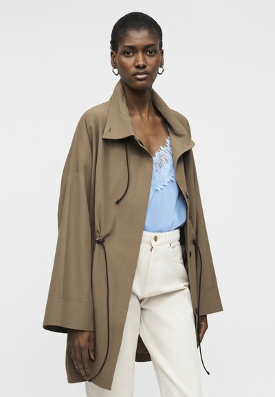 Žena v ľahko voľnej hnedej bunde cez svetlomodrý top s čipkovaným lemom a biele vysokopásové nohavice, stojaca pred jednoduchým pozadím.