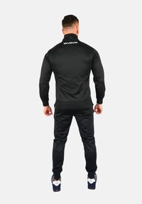 Zwarte sportjacket met een hoge kraag en ribbelmanchetten. Materiaal lijkt glad en aansluitend, met een wit logo op de achterkant. Gecombineerd met zwarte joggers.