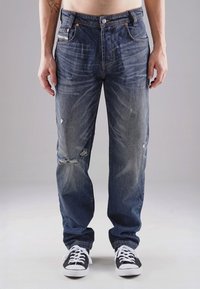Blaue Jeans aus Denim mit lockerer Passform, versehen mit abgenutzten Stellen und ausgeblichenen Flecken. Kombiniert mit schwarz-weißen Sneakern.