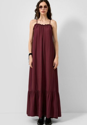 Femme portant des lunettes de soleil rondes et une robe longue sans manches, ample, rouge foncé avec des bretelles fines et des bottines noires.