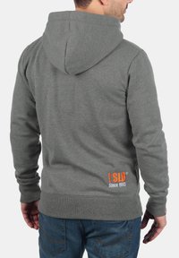 Grijze hoodie met een trekkoord capuchon. De stof is zacht en heeft een comfortabele pasvorm. Heeft een oranje logo en tekst op de onderrug.