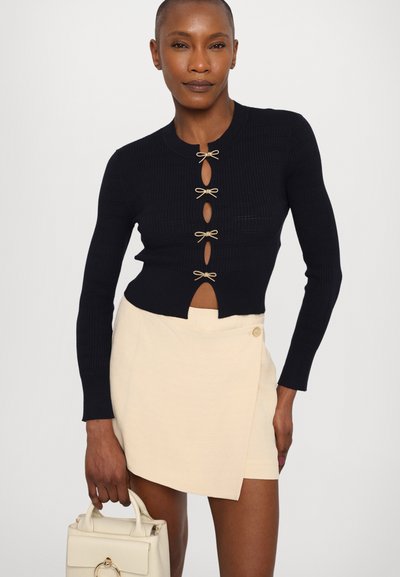 Mornarska rebrasta kardigan sa zlatnim mašnama i otvorima u obliku ključa, uparen sa svijetlom bež suknjom na preklop, držanom malom krem torbicom.